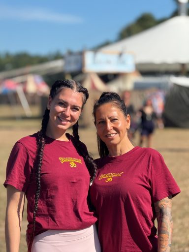 Zwei Frauen in roten T-Shirts lächeln vor einem Zirkuszelt auf einem Festival.