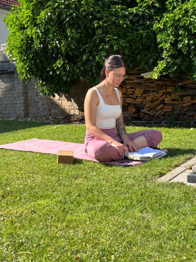 Eine Person sitzt meditierend auf einer Matte im Garten, umgeben von Gras und Pflanzen.