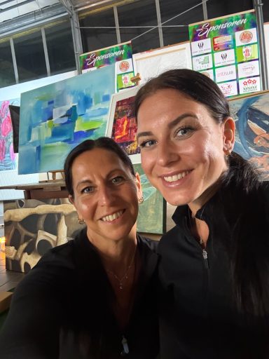 Zwei Frauen in Schwarz lächeln vor bunten Kunstwerken in einem Atelier.