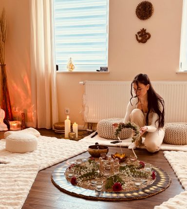 Frau sitzt in einem hellen Raum und gestaltet eine rituelle Anordnung mit Pflanzen und Kerzen.