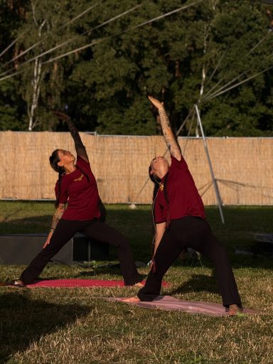 Zwei Personen in roten Shirts üben Yoga in einer grünen Wiese unter blauem Himmel.